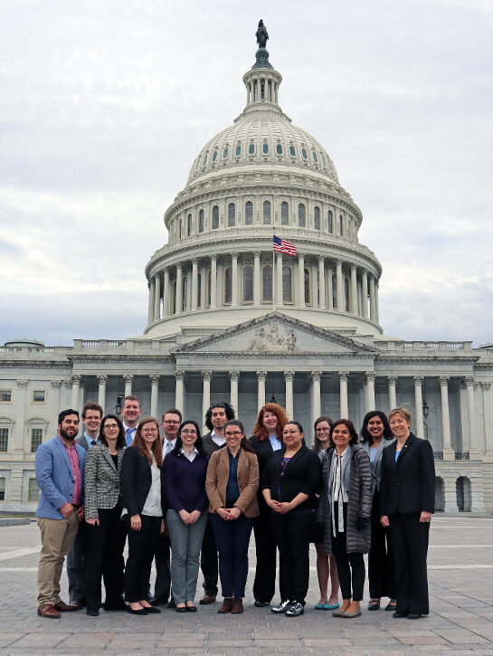 A group of people stand shoulder-to-shoulder in a group photo in frone of the U.S. Capitol dome in the background.