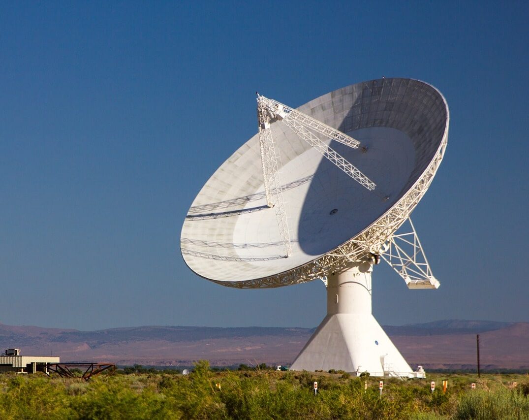 A large dish-shaped radio telescope antenna is seen against a blue daytime sky. The sun is casting shadows on its surface. Vegetation appears in the foreground.