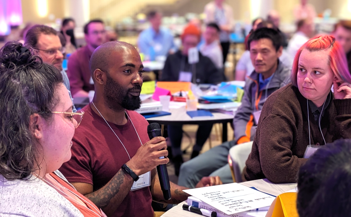 A group of people are in a discussion session with one person holding a microphone and speaking to others