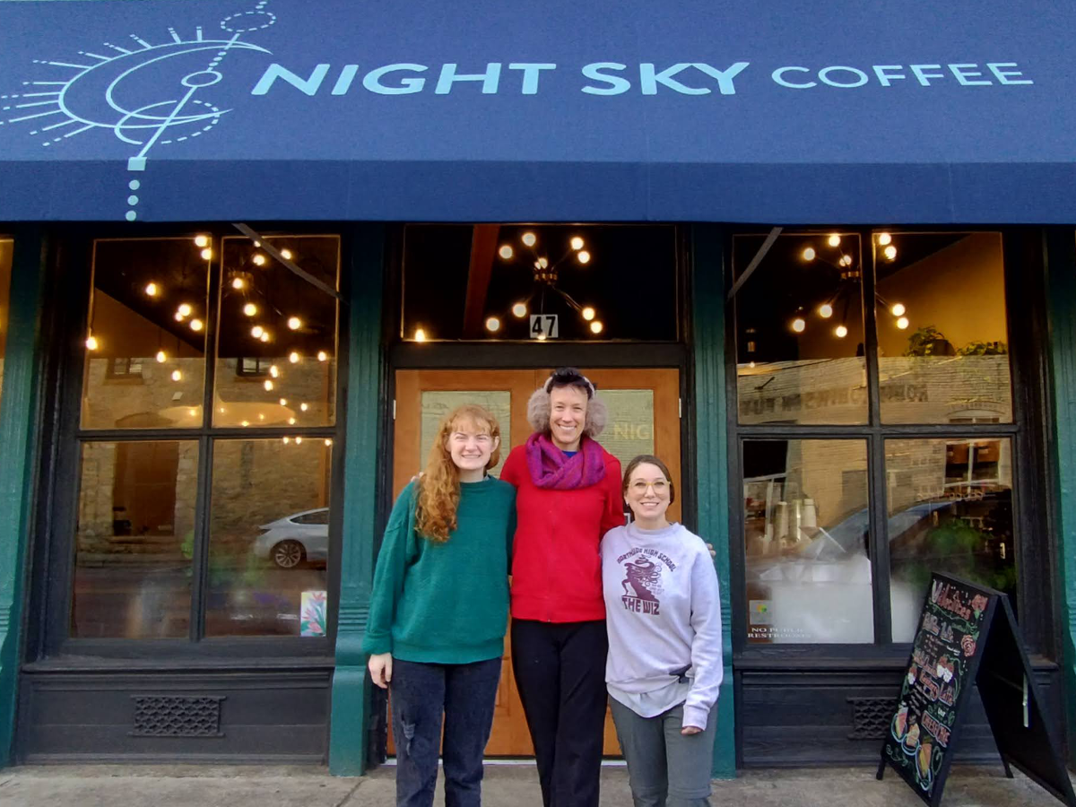 Three people stand before a storefront. A blue awning is above them printed with the words "Night Sky Coffee" in a lighter shade of blue. Lights are visible through the windows of the store. An A-frame sign with menu items appears at lower-left.