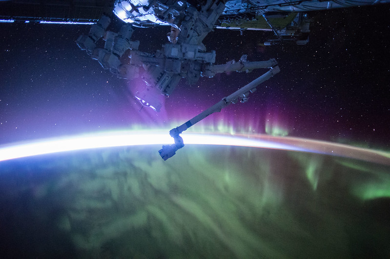 A view of space over the limb of the Earth, which takes up the lower half of the image. Red and green aurorae hover over the planet. The Sun is rising on the horizon. Stars appear above the Earth along with part of the International Space Station.