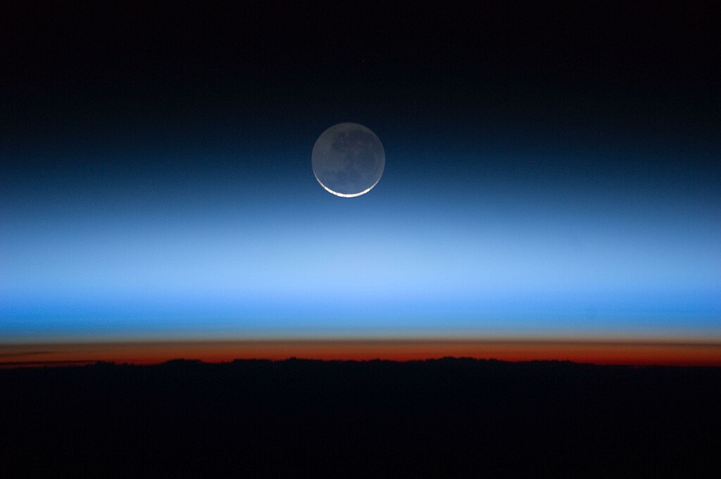 A crescent moon appears suspended in the blackness of space above the Earth's limb. The atmosphere is a translucent bluish haze, going to dark red near the horizon.