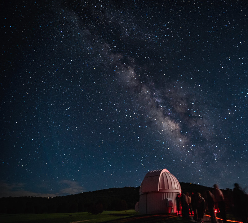 A starry night sky takes up most of the frame. The Milky Way arches from the horizon at lower right to the overhead point at top-center. In the foreground at right is a small observatory dome with people standing in line waiting to look through the telescope. Mountains are on the horizon.