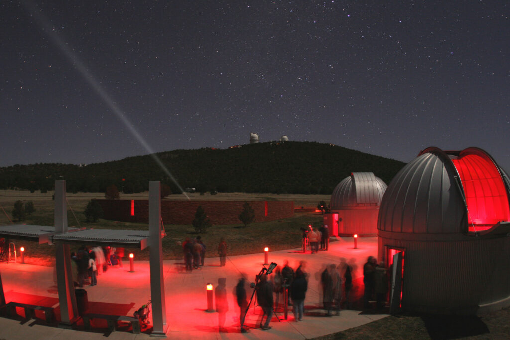 A group of people stand outside an observatory dome under a dark night sky. The scene is dominated by illumination from red lights. In the distance at center, two telscope domes sit atop a mountain.