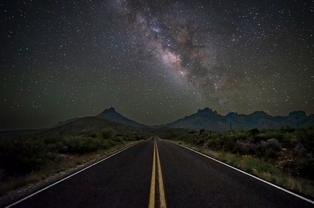 A night scene in which an asphalt road disappears into the center of a desert landscape. The Milky Way and numerous stars appear in the sky overhead. There are dark mountains on the horizon.