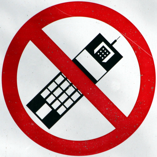 A black and white cartoon representation of a mobile phone tilted at a 45 degree angle is covered by the red circle-and-sash symbol meaning that phones are prohibited.