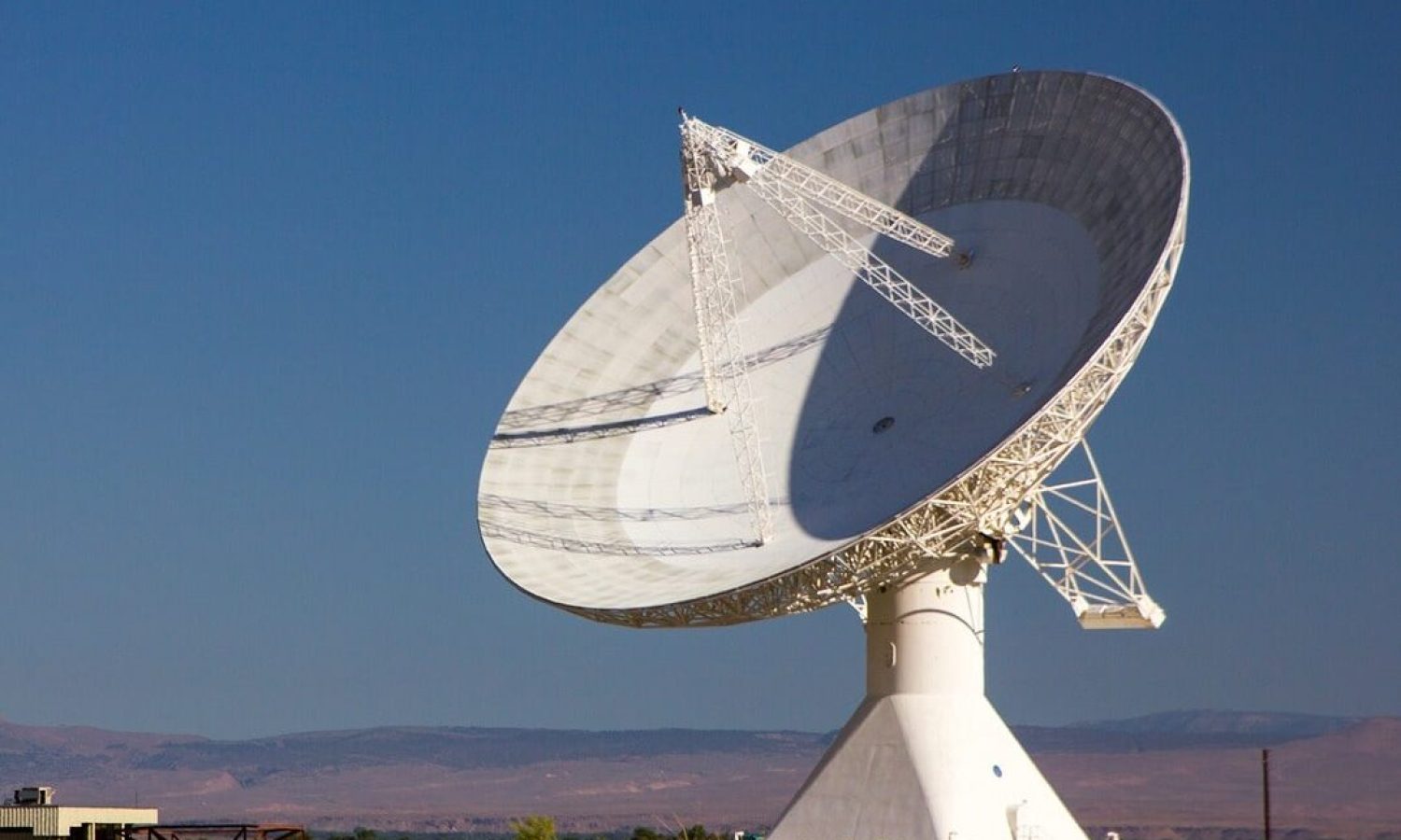 A large dish-shaped radio telescope antenna is seen against a blue daytime sky. The sun is casting shadows on its surface. Vegetation appears in the foreground.